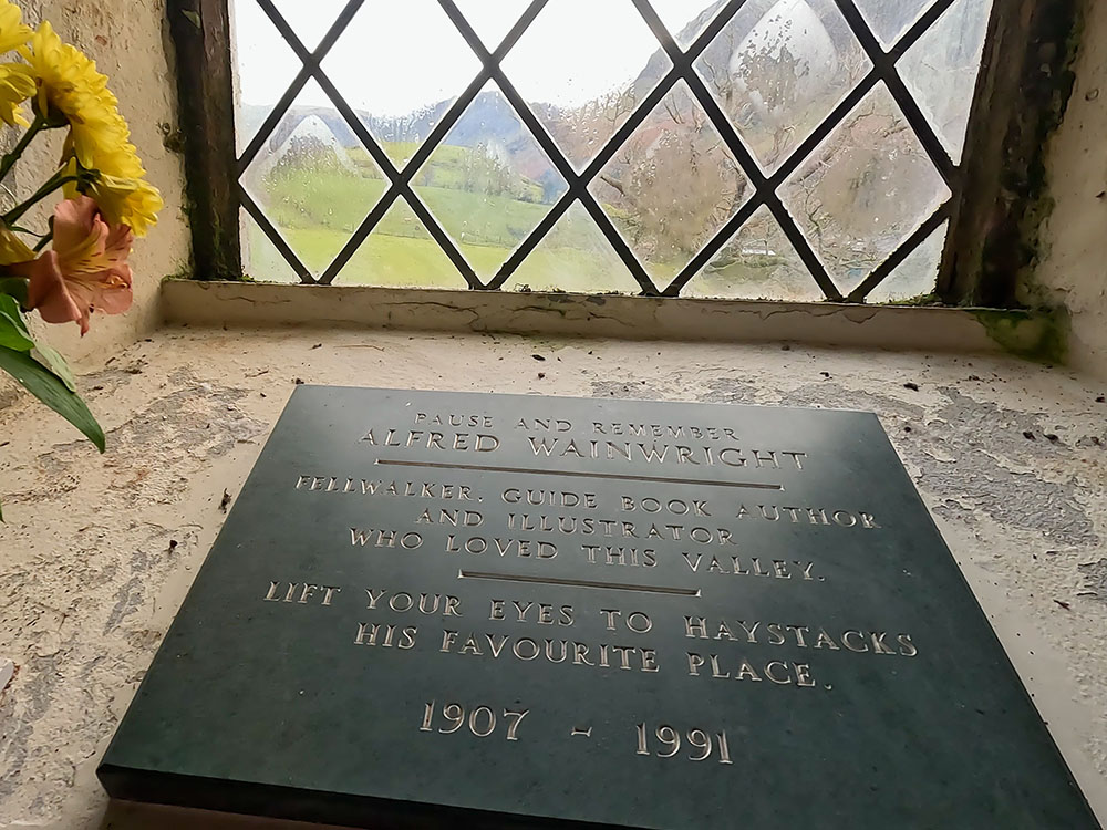 A stone plaque commemorating Wainwright in the church at Buttermere. Through the window on the horizon is Haystacks.