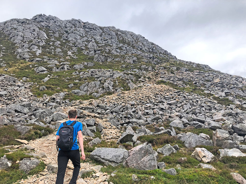 Photo of an adult walker approaching rocky hill terrain on a UK route under cloudy skies.
