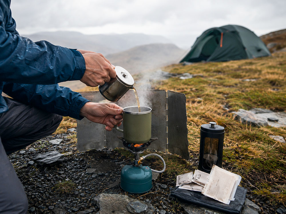 UK hill camping scene showing a safe stove and wind shield coffee setup in damp, windy weather with a tent in the background and steam rising from a mug.
