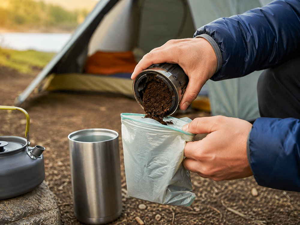 Close-up of used coffee grounds being packed into a sealable waste bag at a tidy campsite, showing responsible outdoor coffee clean-up with a mug and kettle nearby.