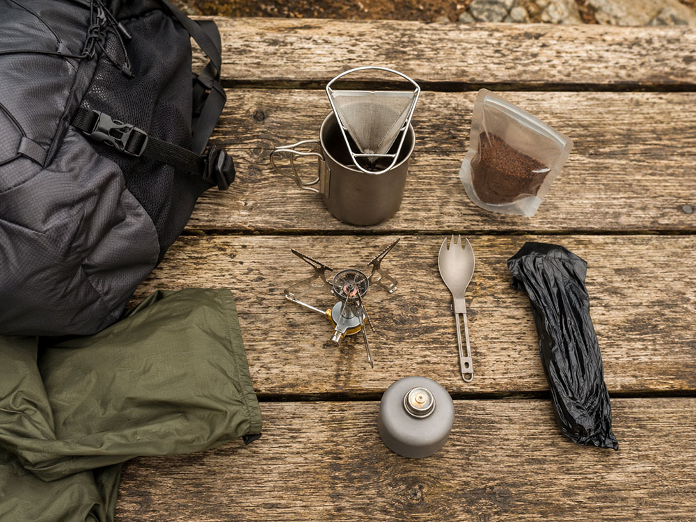 Lightweight backpacking coffee kit laid flat out with mesh pour-over filter, titanium-style mug, stove, fuel canister, ground coffee, spork, waste bag, jacket and rucksack.