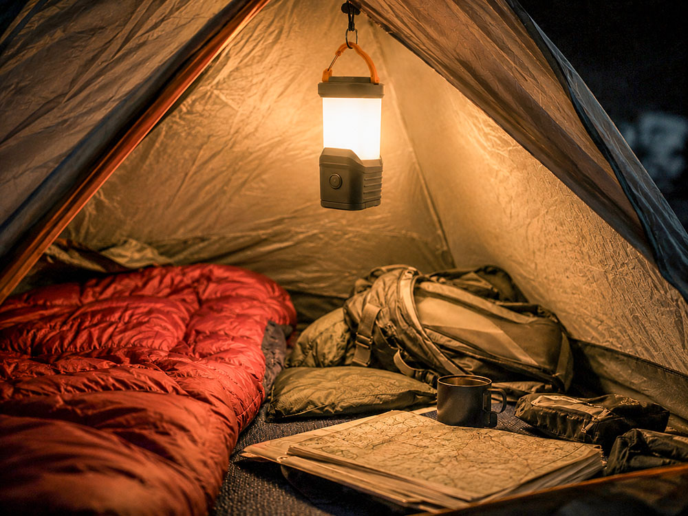 Modern camping lantern hanging inside a tent, lighting a sleeping bag, rucksack, walking map and camping kit at night.