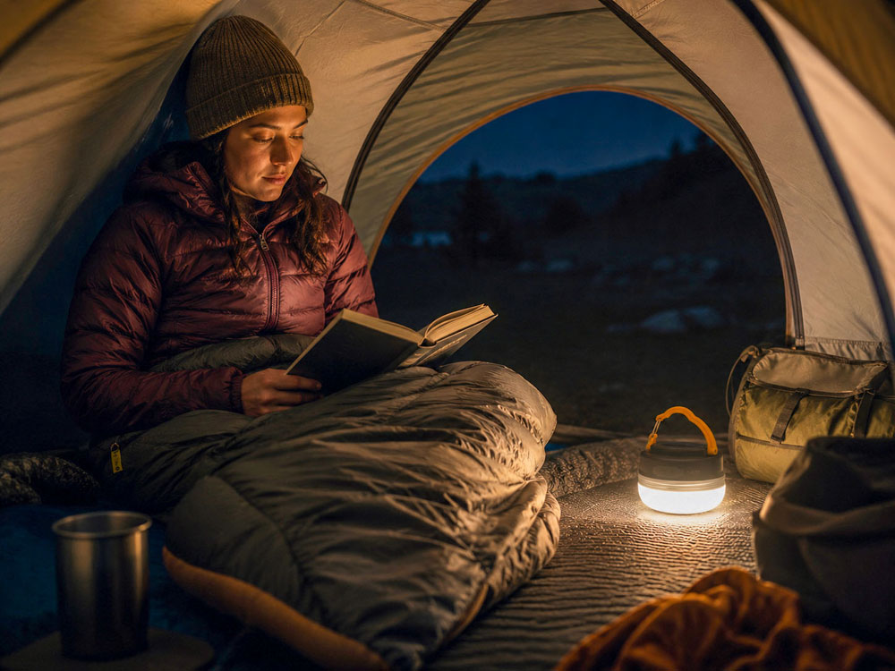 Camper reading inside a tent at night using a compact modern camping lantern for warm, comfortable light.