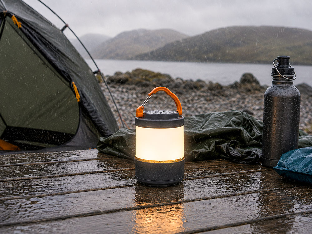 Modern water-resistant camping lantern glowing on a wet table beside a tent during rainy UK camping weather.