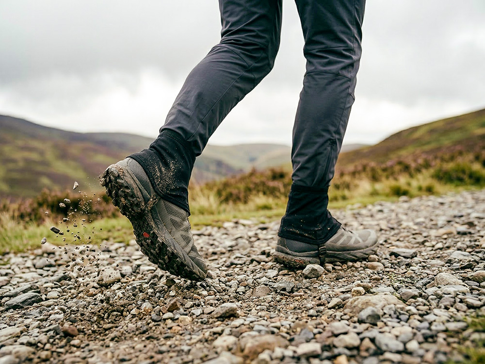 A trail runner on a stony fell path wearing low-profile ankle gaiters and running shoes