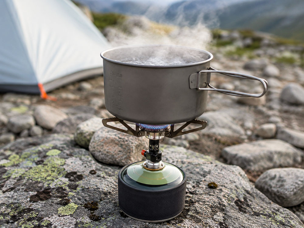 Close-up of a lightweight camping stove boiling water in a metal pot on rocky ground, showing practical outdoor cooking performance.