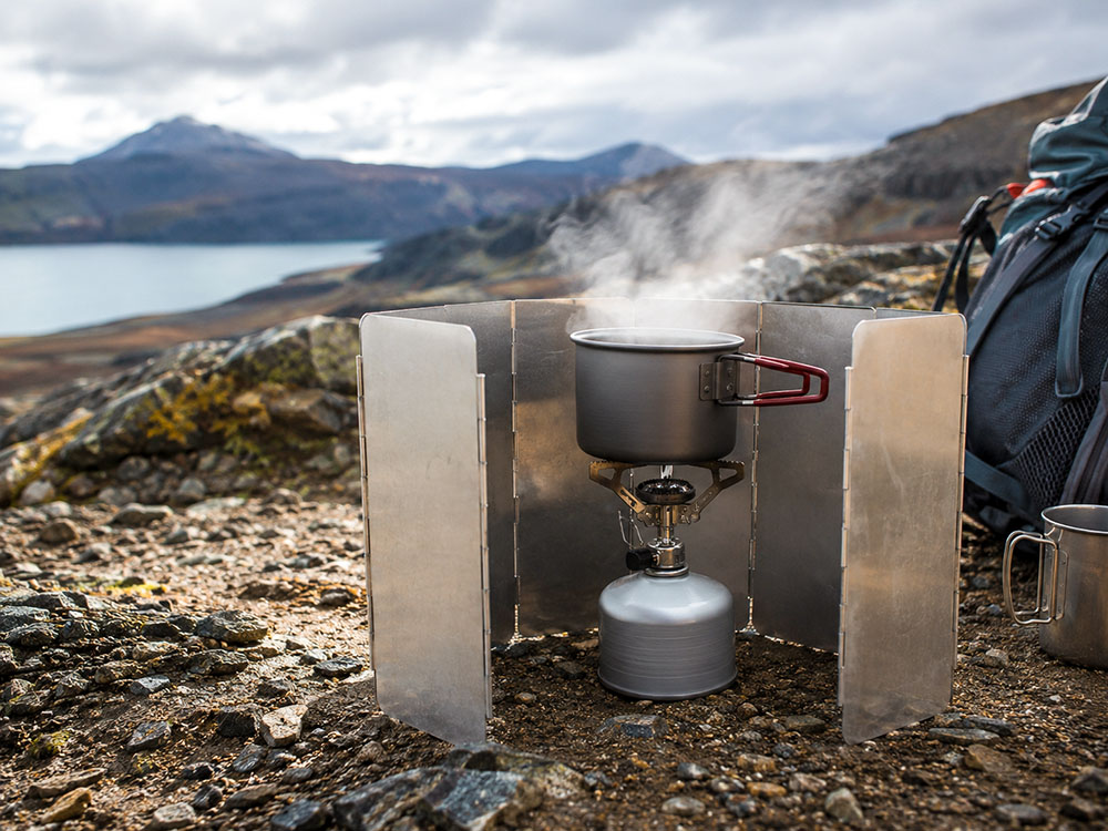 Lightweight camping stove boiling water behind a windshield in windy mountain conditions, showing safer stove use outdoors.