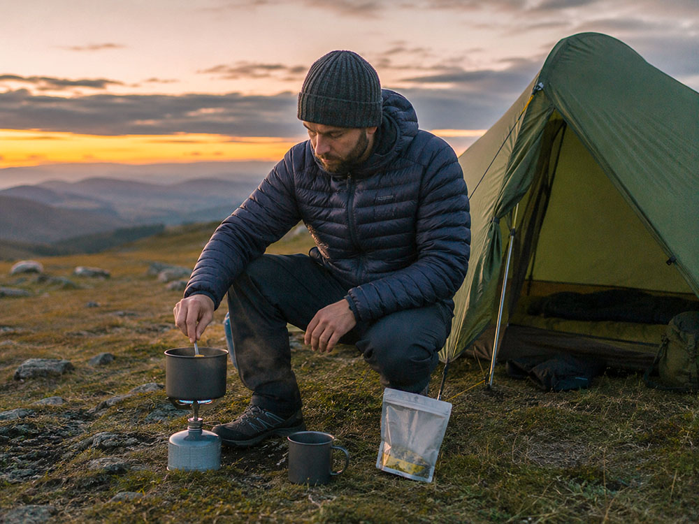Hiker cooking beside a small backpacking tent at dusk with a compact lightweight camping stove, mug and outdoor meal pouch.