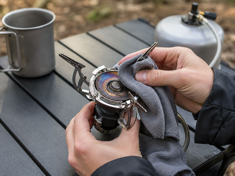 Hands cleaning a lightweight camping stove burner with a cloth after use on an outdoor table, showing practical stove maintenance.