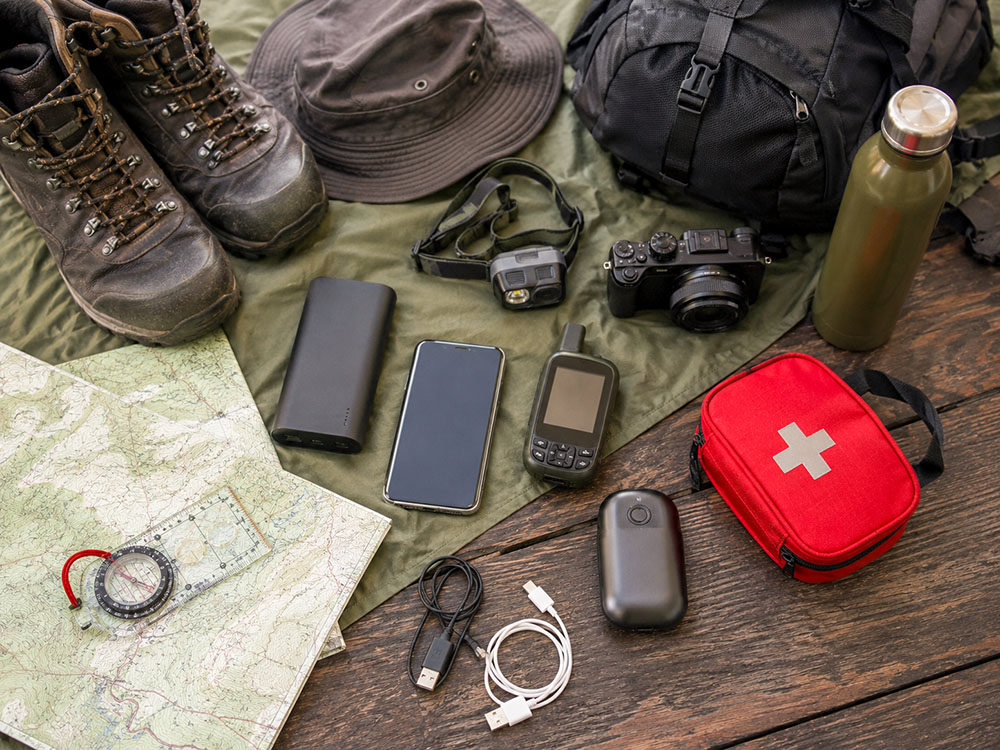 Flat lay of camping and walking gear including a power bank, GPS, head torch, phone, hand warmer, camera, map and compass.