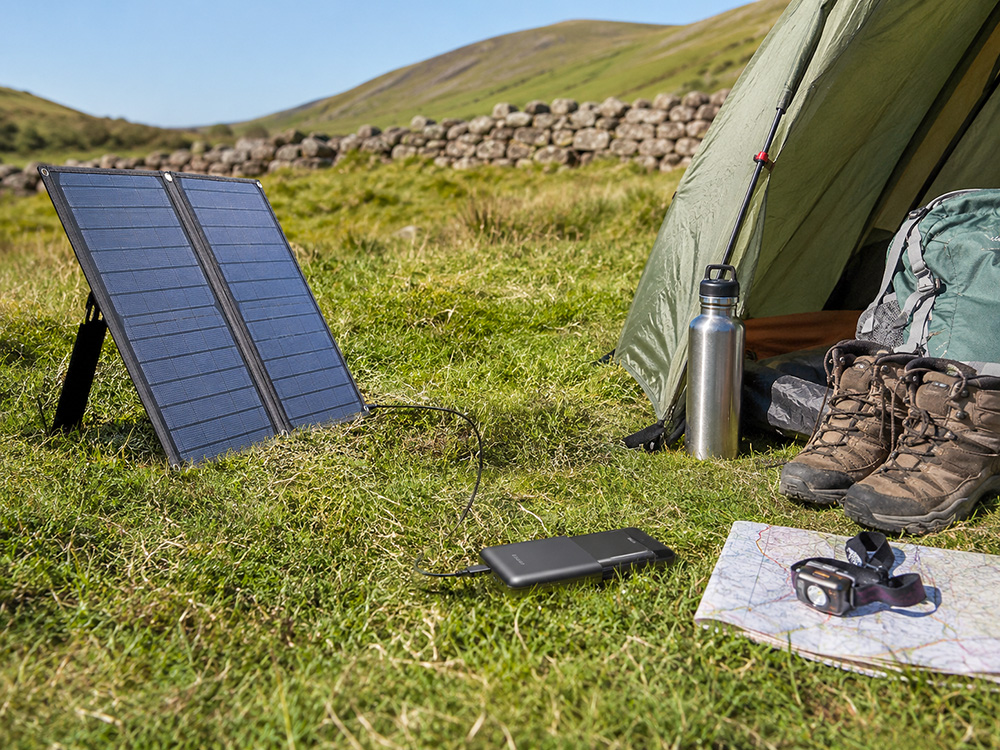 Foldable solar panel charging a power bank at a countryside campsite with walking boots, tent and outdoor gear nearby.