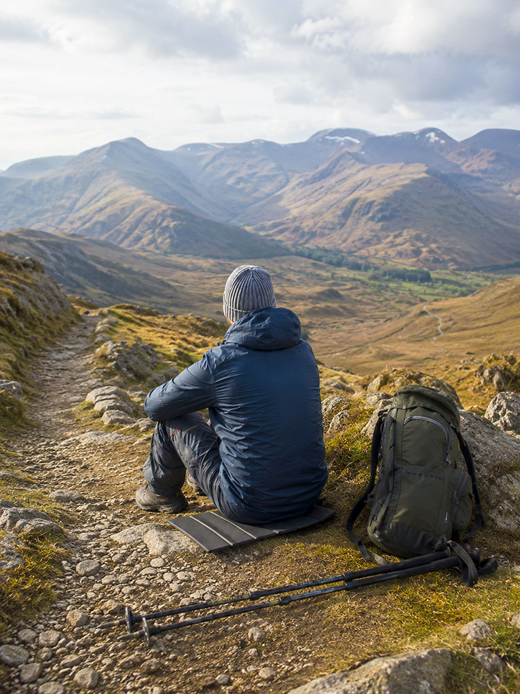 Long-distance walker taking a short trail break on a lightweight sit mat, with rucksack, walking poles and open UK hill path nearby.