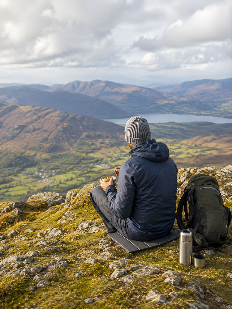 Hill walker having lunch on a compact sit mat during a UK mountain walk, with rucksack, flask and wide upland views.