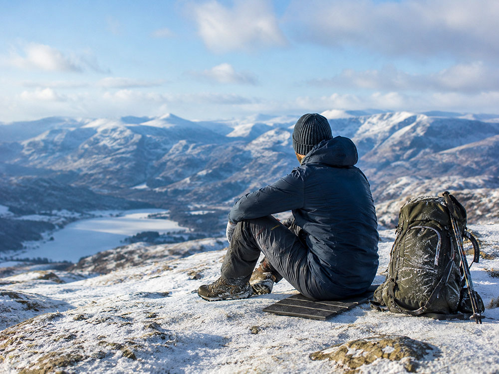 Winter walker sitting on an insulated sit mat on snowy or frosty ground, with rucksack and cold UK mountain scenery behind.