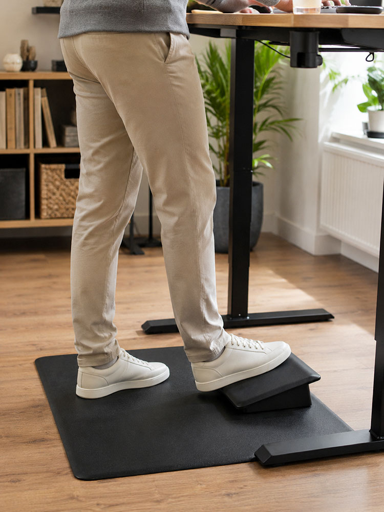 Person using an anti-fatigue mat and footrest step at a standing desk for standing comfort and posture variation.