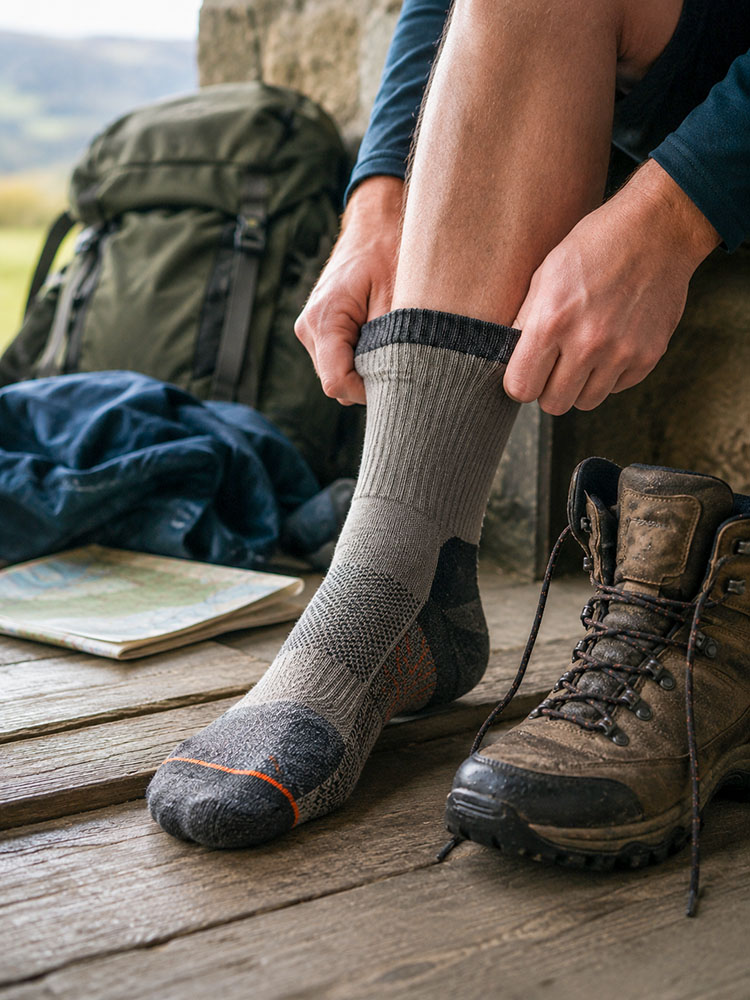 Walker pulling up a smooth grey walking sock before putting on a muddy walking boot, with rucksack, map and outdoor kit nearby.