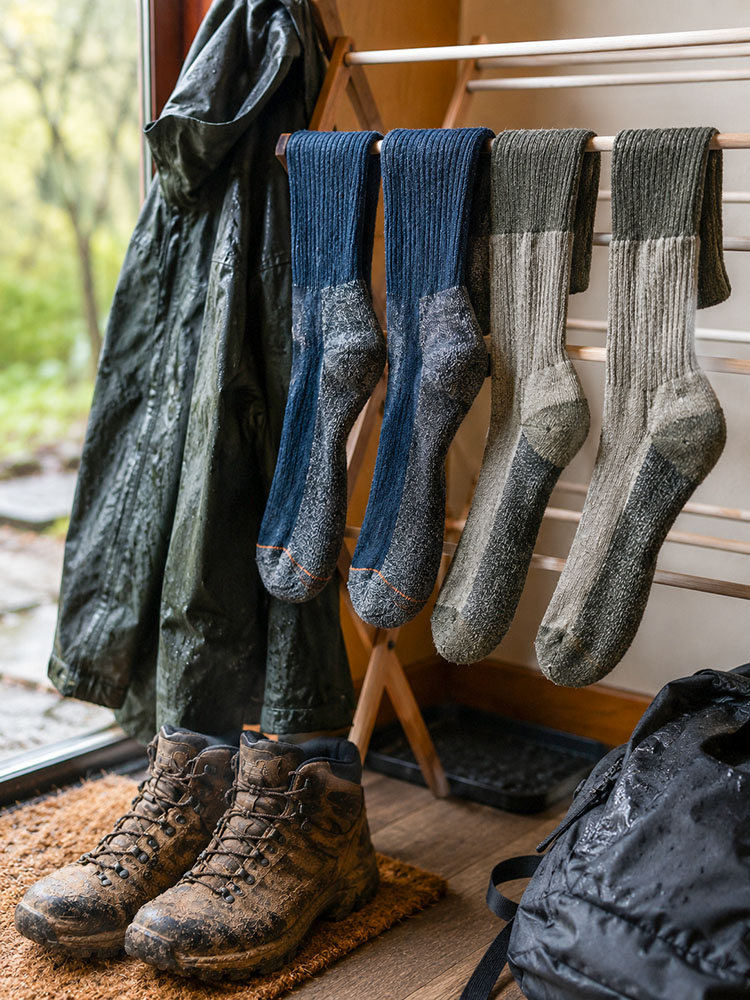 Walking socks drying on an indoor rack beside muddy boots, a wet waterproof jacket and a rucksack after a rainy walk.