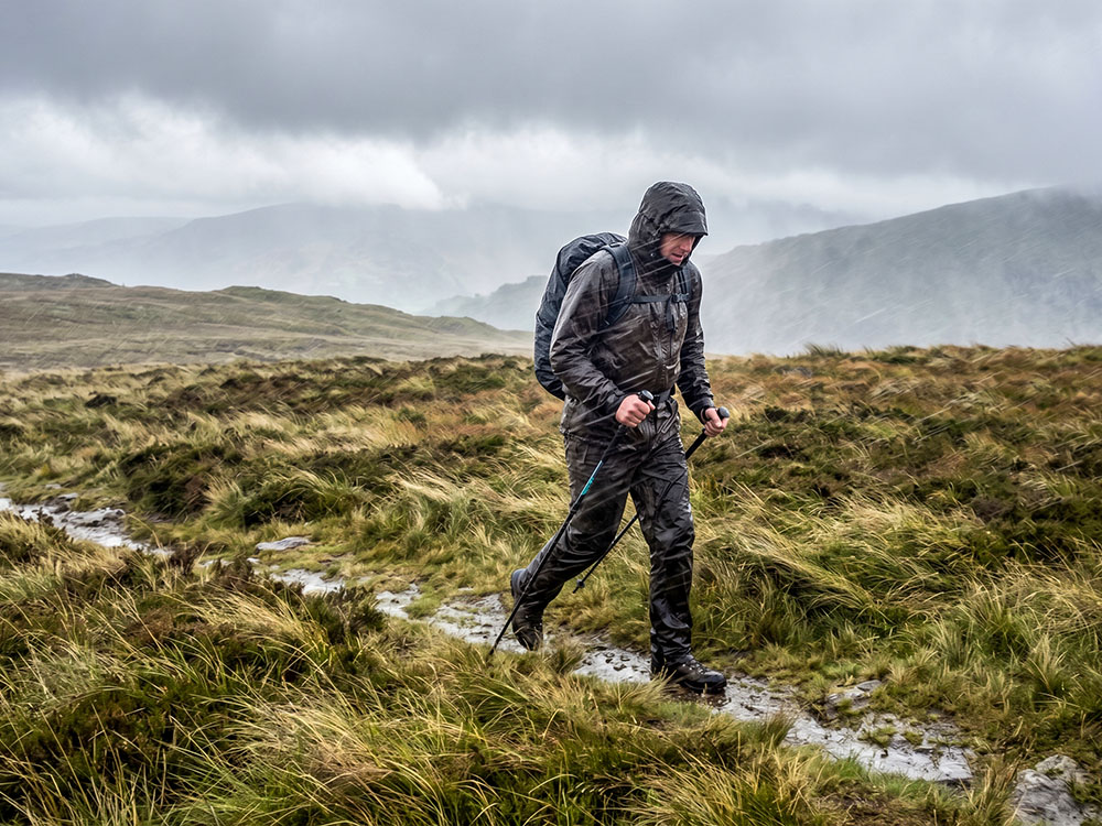 Hillwalker crossing exposed British moorland in wind-driven rain wearing a waterproof jacket and waterproof trousers